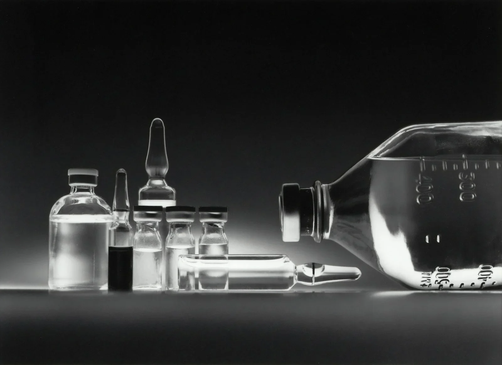 Glass medical vials and a large intravenous fluid bottle on a dark surface